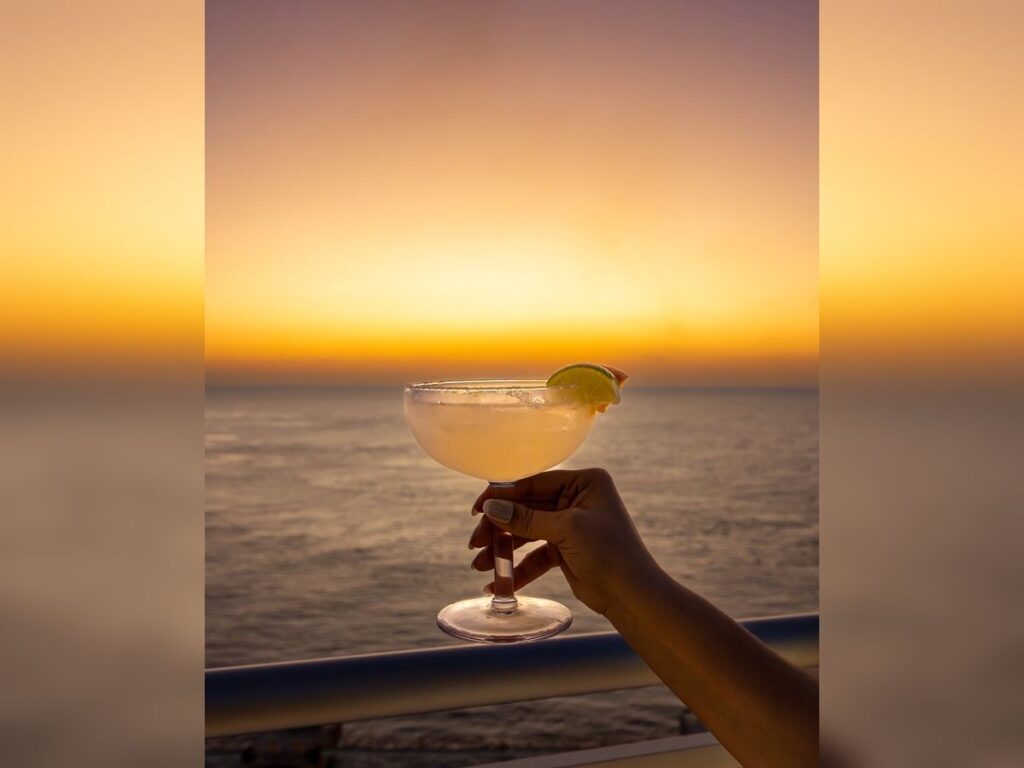 Hand holds a margarita with a salted rim and lime wedge on a cruise ship balcony at sunset over the ocean.