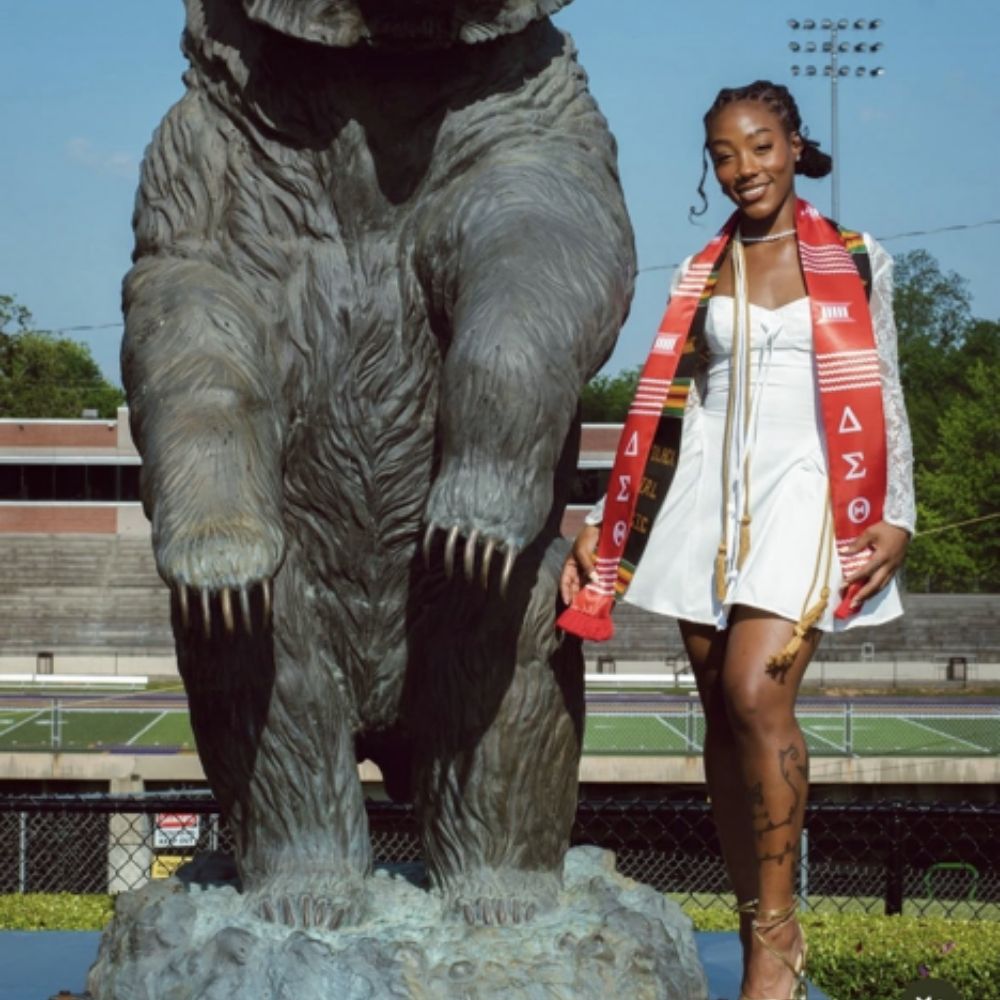 A young woman, Hannah Smith, in a white graduation outfit and red stole poses beside a large bear statue on a sports field.