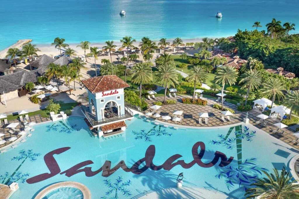 Aerial view of a Sandals resort with a large swimming pool, palm trees, and a white-sand Caribbean beach.