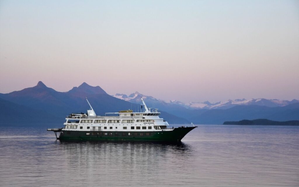 Small Uncruise expedition cruise ship sailing on calm water at dusk with mountain peaks in the background