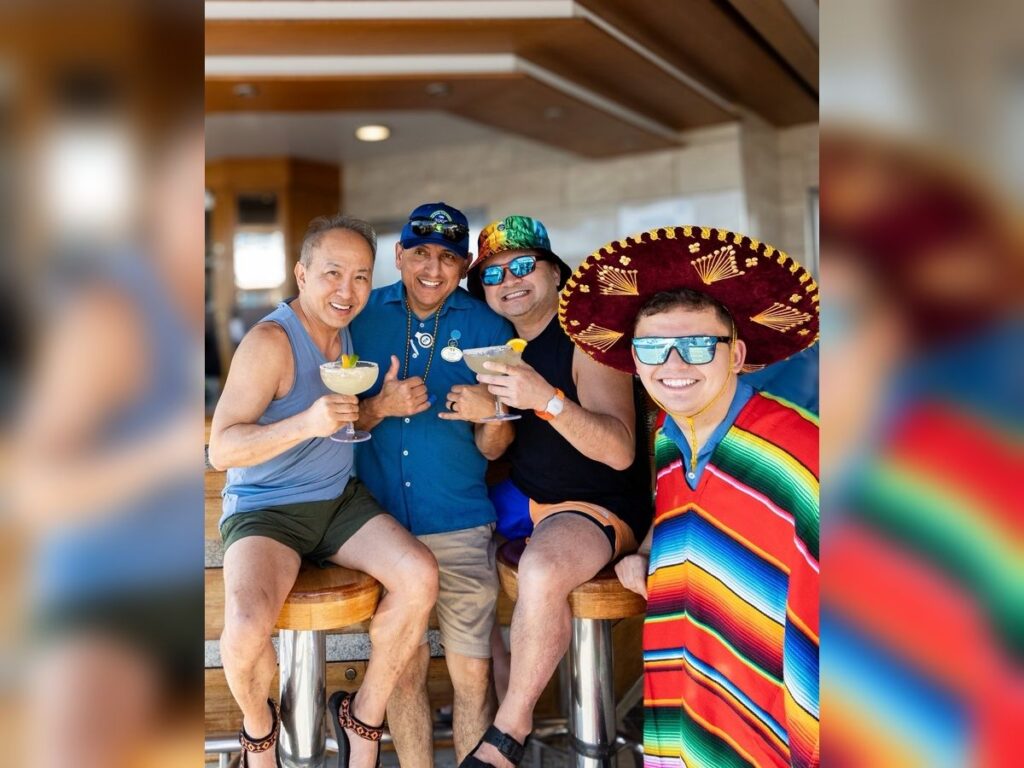 Cruise guests pose together with crew member at a ship bar holding margaritas, with one wearing a sombrero and a colorful serape.
