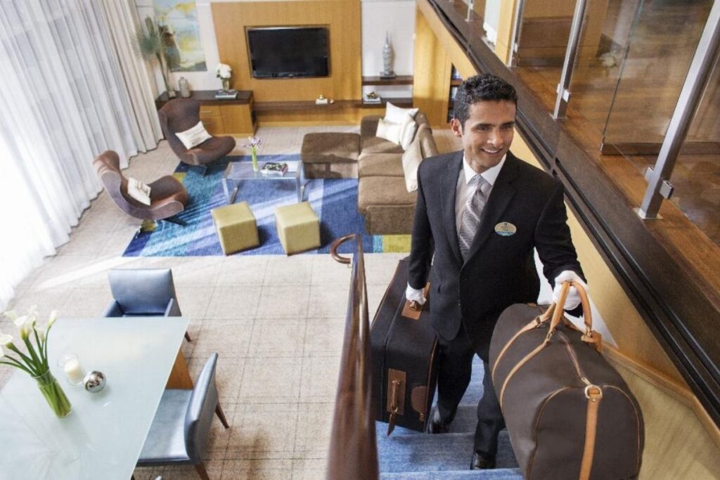 A suited cruise butler walks up the stairs of a spacious two-level suite carrying luggage, with a large living area, dining table, and floor-to-ceiling curtains visible behind him.
