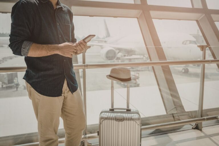 Traveler holding a phone beside a silver suitcase in an airport terminal, with airplanes visible through large windows in the background.