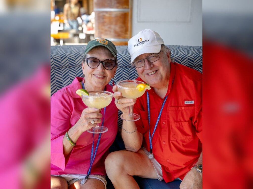An older couple smiles on a cruise ship lounge seat while holding salt-rimmed margaritas garnished with lime and lemon.