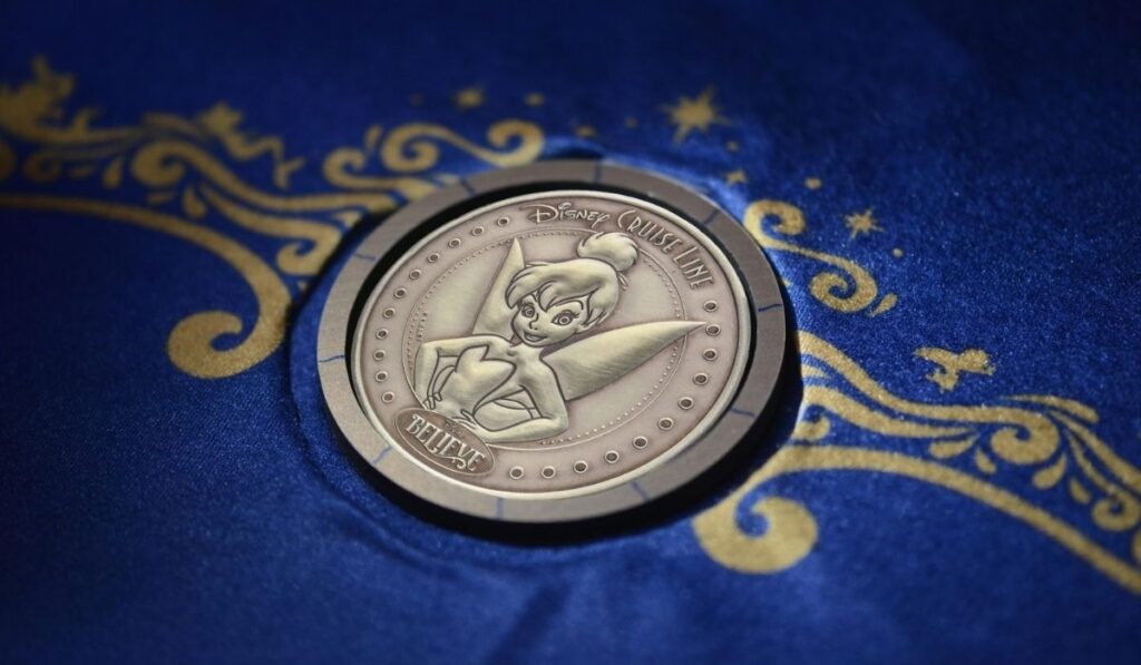 Close-up of the Disney Believe commemorative keel-laying coin featuring Tinker Bell, resting on blue fabric with gold decorative patterns.