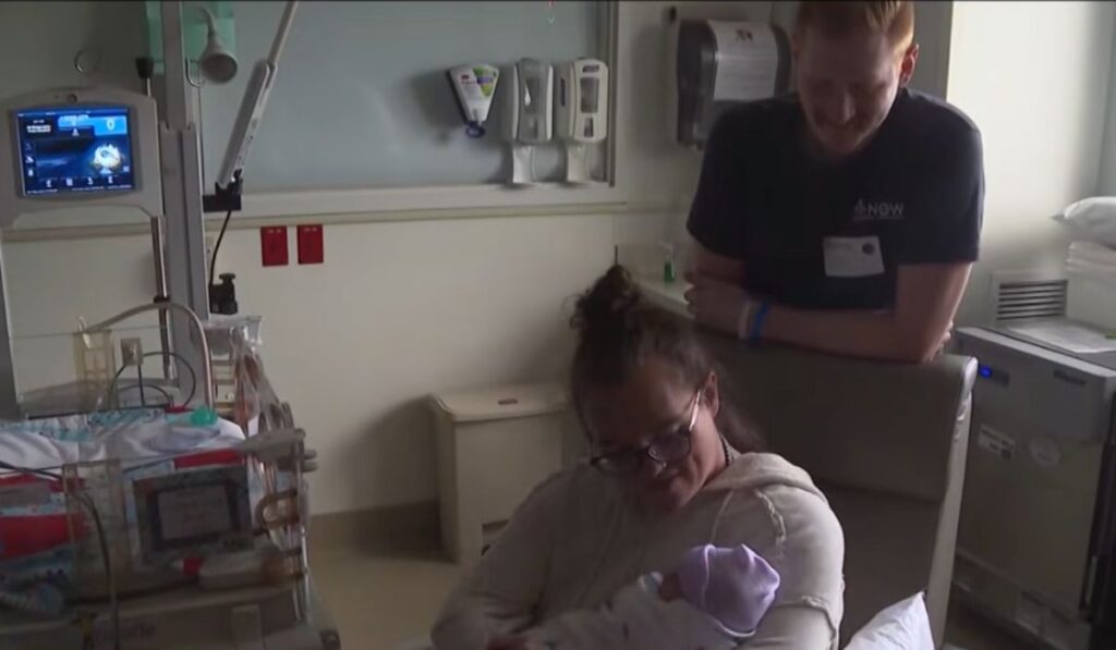A mother holding her newborn baby in a hospital room while the baby’s father stands behind her.