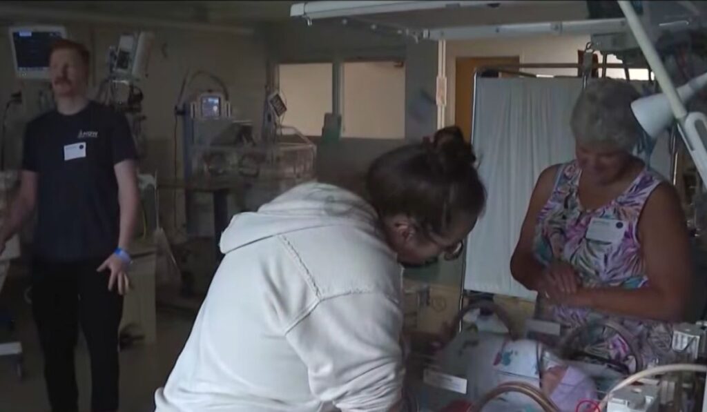 A new mother leaning over a hospital bassinet while a nurse and the baby’s father stand nearby.