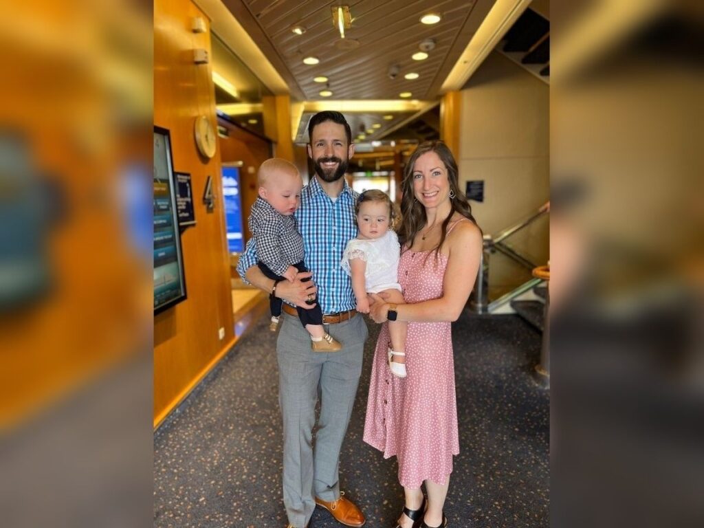 Family of four poses in a cruise ship hallway, with a dad in a blue check collared shirt with blue slacks and dress shoes holding a toddler boy and a mom in a pink polka-dot dress and wedges holding a toddler girl.