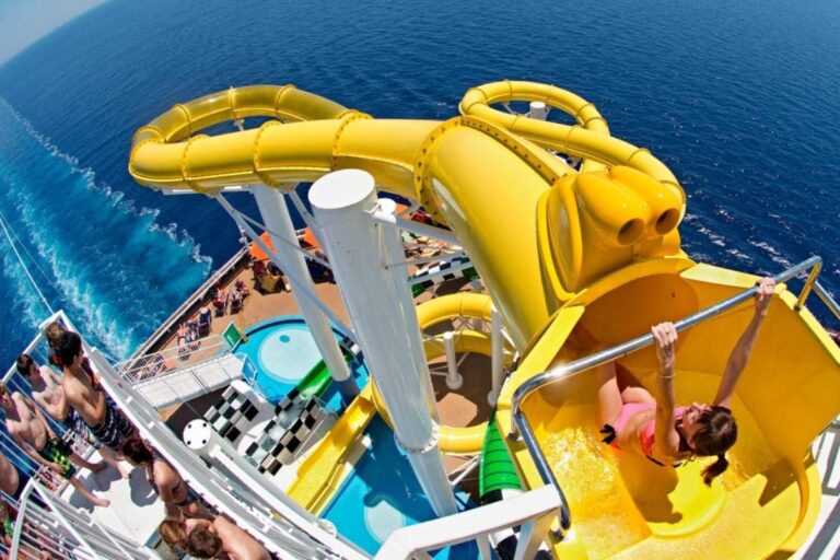 Woman starts down a yellow waterslide on Carnival Sunshine cruise ship, with ocean views and guests watching nearby.