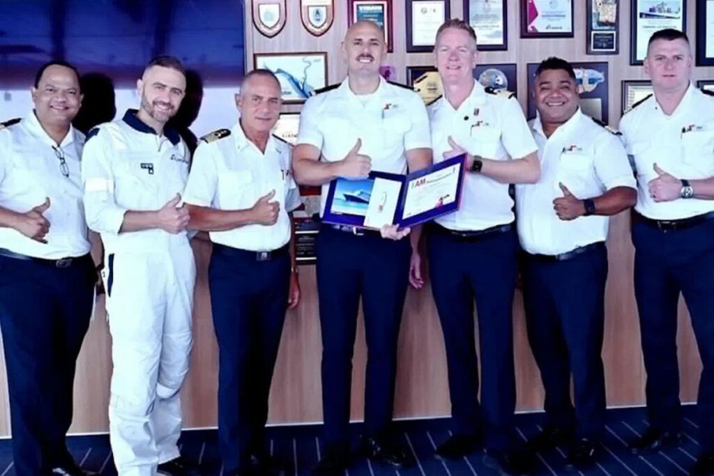 Six Carnival crew members in white uniforms pose together onboard as one crew member holds a Hero award certificate after being recognized for saving a choking baby on Carnival Celebration.