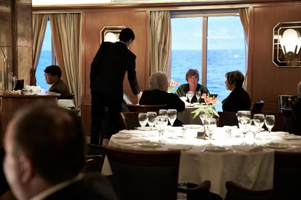 Cruise ship dining room with a server assisting guests at tables beside large windows overlooking the ocean.