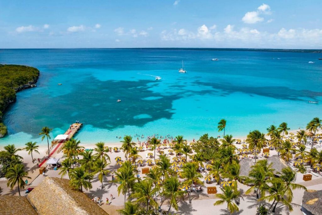 Aerial view of Catalina Island beach with rows of yellow-and-white umbrellas, palm trees, a small pier, swimmers, and boats on bright turquoise water