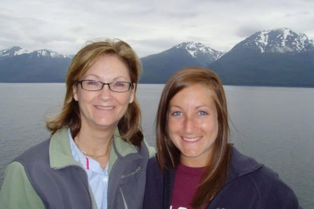 Two women smiling for a photo in front of calm water and snow-capped mountains in Alaska under an overcast sky.