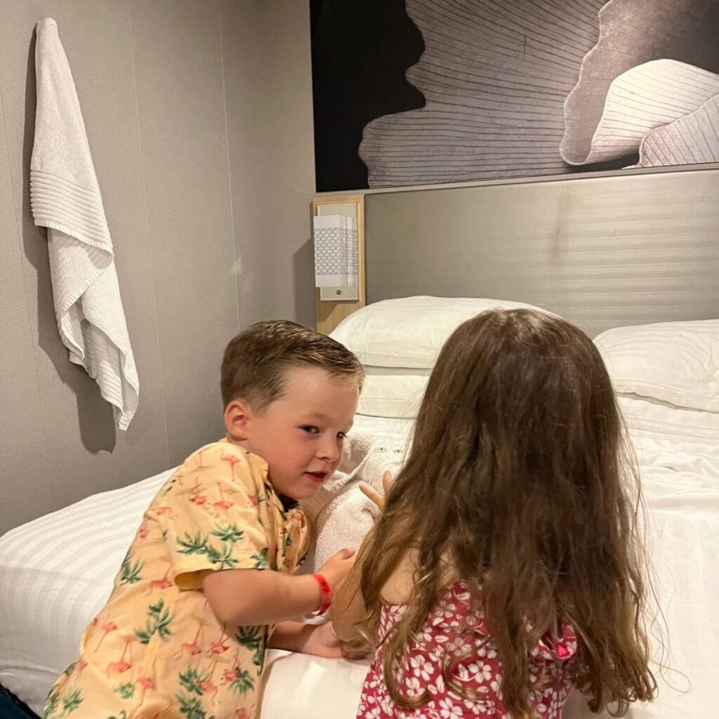 Two children playing on a white bed inside a cruise ship cabin, with white pillows, towels hanging on the wall, and a bedside light visible behind them.