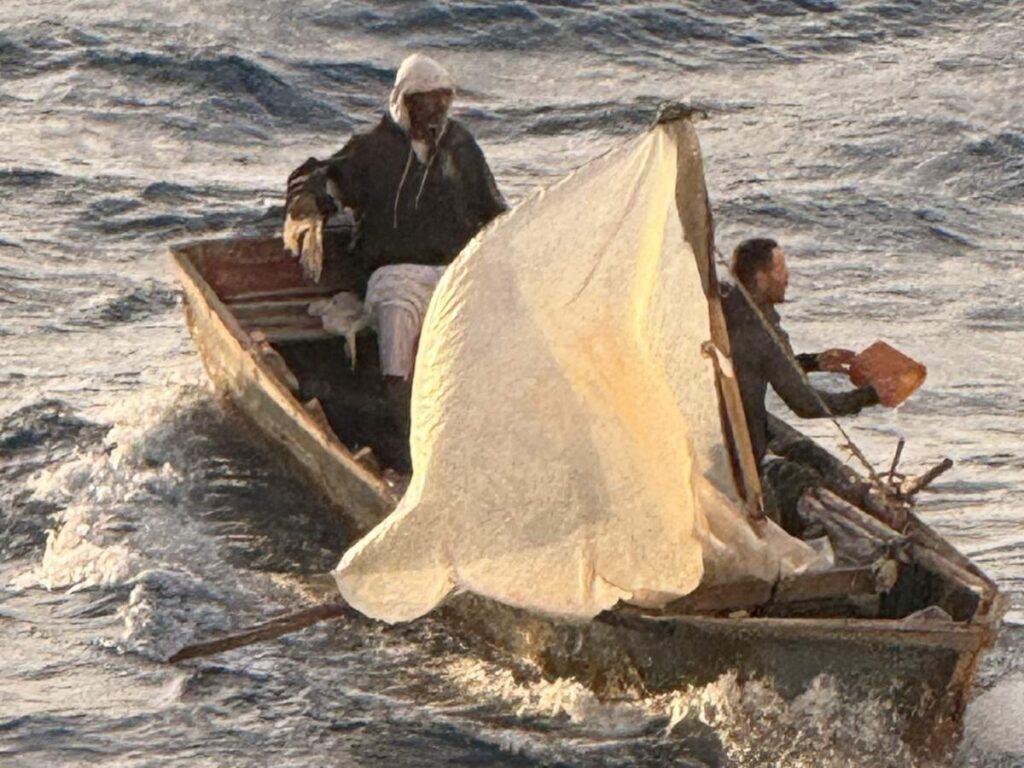 Close-up of a small wooden boat with a makeshift white sail as two people brace against rough seas and have bucket in hand bailing out water during a rescue at sea.