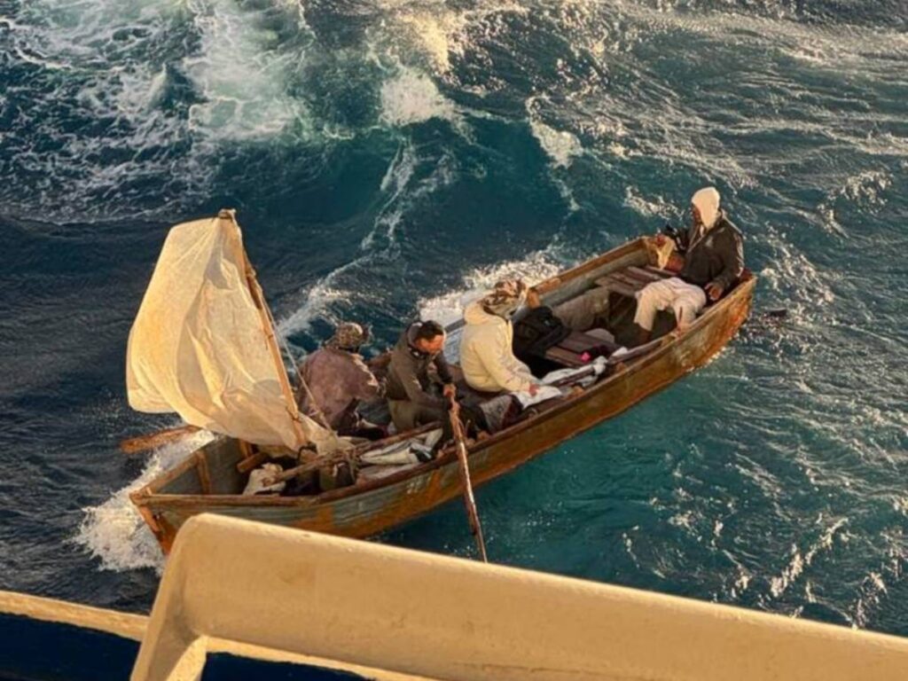 Four people sit in a small wooden boat with a makeshift white sail as it drifts in choppy water alongside the Regal Princess cruise ship during a rescue.