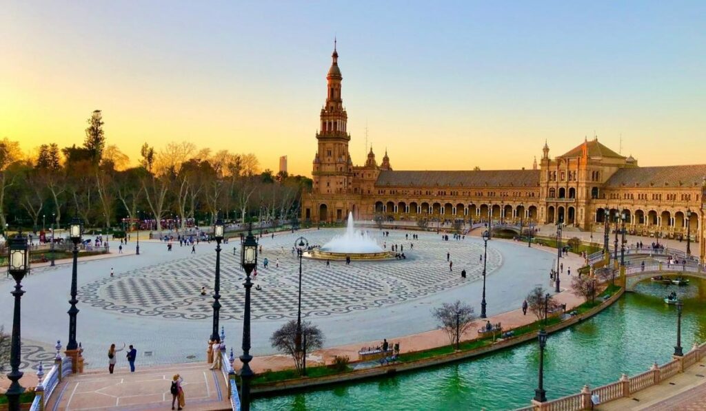 Aerial view of Sevilla's Plaza de España at sunset, featuring the iconic tiled buildings, central fountain, moat, and iconic tower in warm golden light.