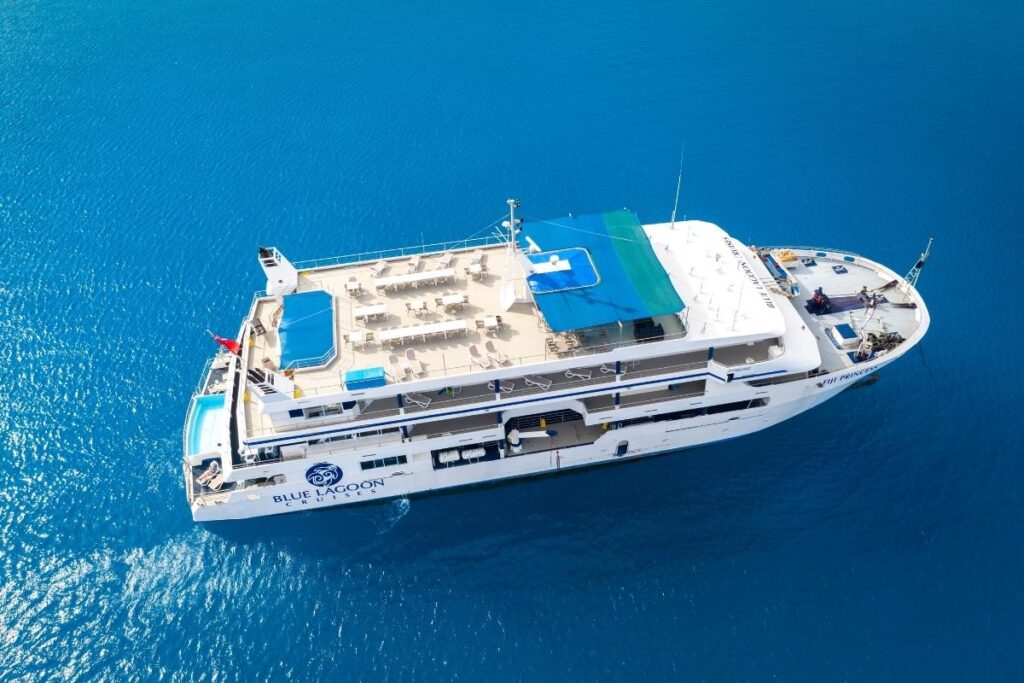 Aerial view of the Fiji Princess cruise ship sailing in bright blue water, with open deck seating, shaded canopy areas, and Blue Lagoon Cruises branding visible on the side.