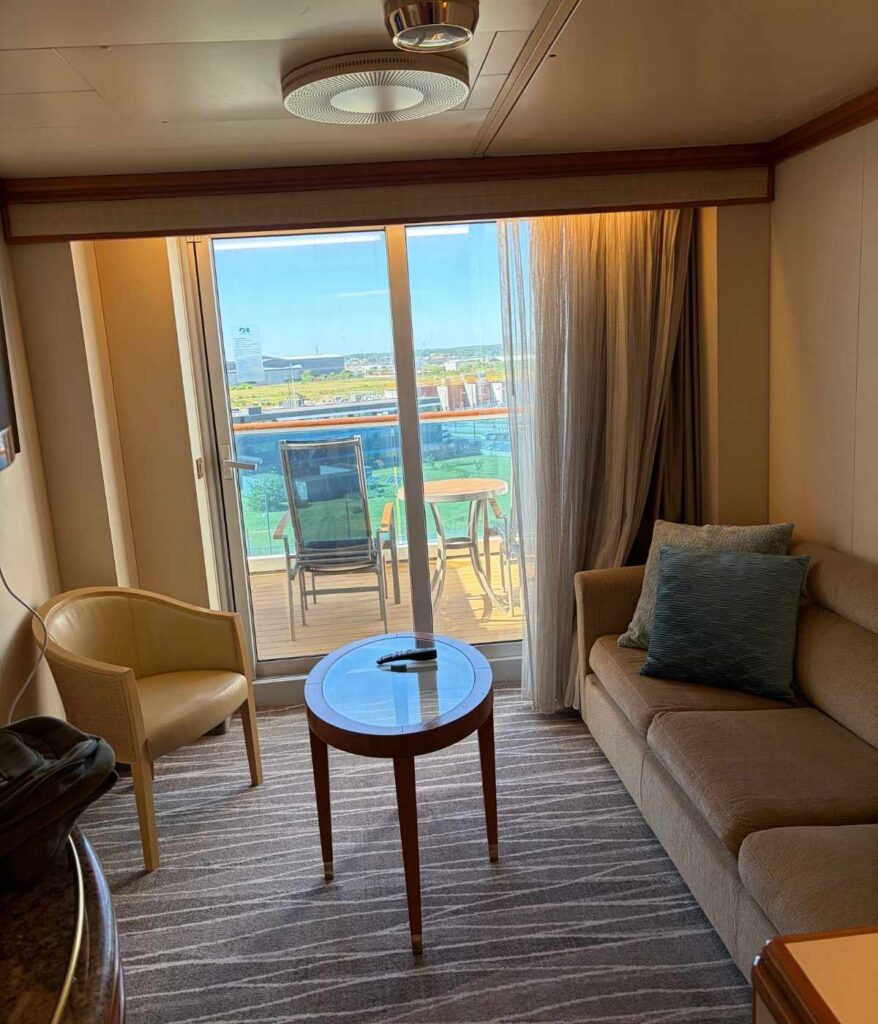 Cruise ship balcony cabin with a sofa, armchair, glass coffee table, and sliding doors opening to a private balcony with chairs and a table.