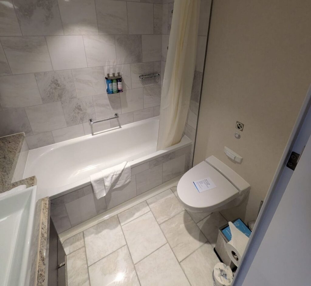 Cruise ship cabin bathroom with a white bathtub, shower curtain, toilet, tiled walls, sink, toiletries, and folded towels.