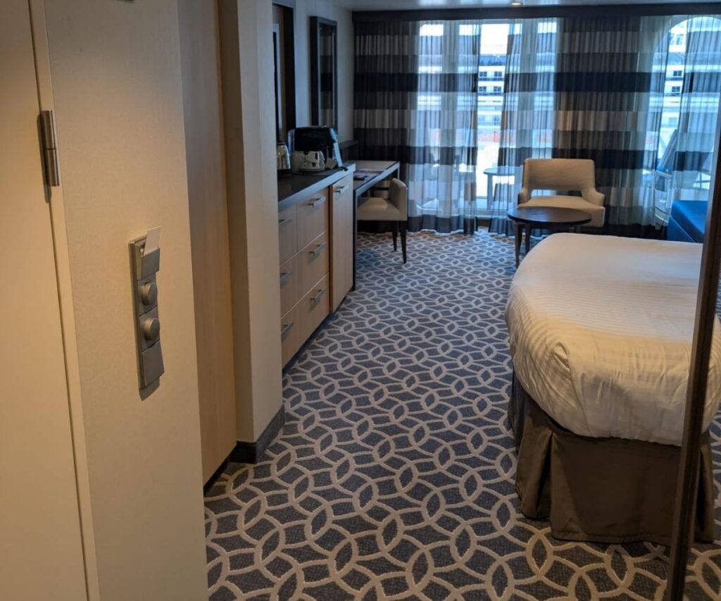 Cruise ship balcony cabin with a queen bed, patterned carpet, desk and drawers, armchair, coffee table, and sliding glass doors leading to a private balcony.