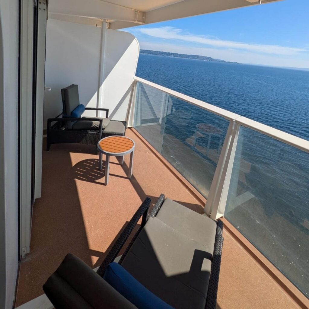 Cruise ship balcony cabin with a queen bed, patterned carpet, desk and drawers, armchair, coffee table, and sliding glass doors leading to a private balcony. Cruise ship balcony with two reclining chairs and a small round table, glass railing, and wide ocean views under a clear blue sky.