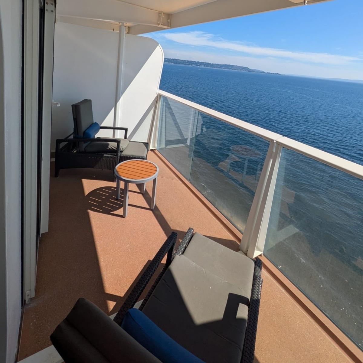 Cruise ship balcony cabin with a queen bed, patterned carpet, desk and drawers, armchair, coffee table, and sliding glass doors leading to a private balcony.Cruise ship balcony with two reclining chairs and a small round table, glass railing, and wide ocean views under a clear blue sky.