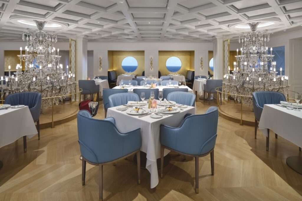 Elegant interior of Le Bistro on Norwegian Viva, with crystal chandeliers, white tablecloth dining tables, blue chairs, and round ocean-view windows.