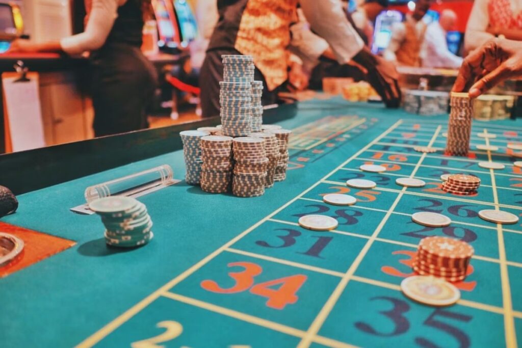 Stacks of casino chips on a roulette table with players and dealers blurred in the background.