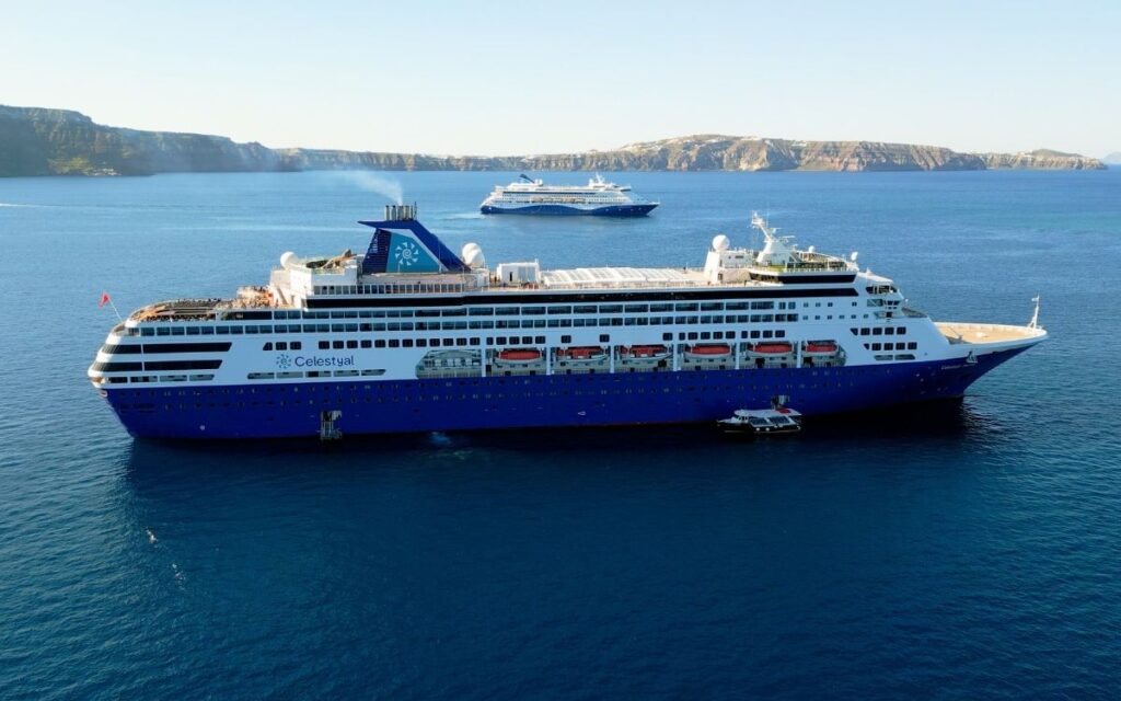 Celestyal Discovery cruise ship in calm blue water with Celestyal Journey cruise ship in the distance near a rocky coastline.