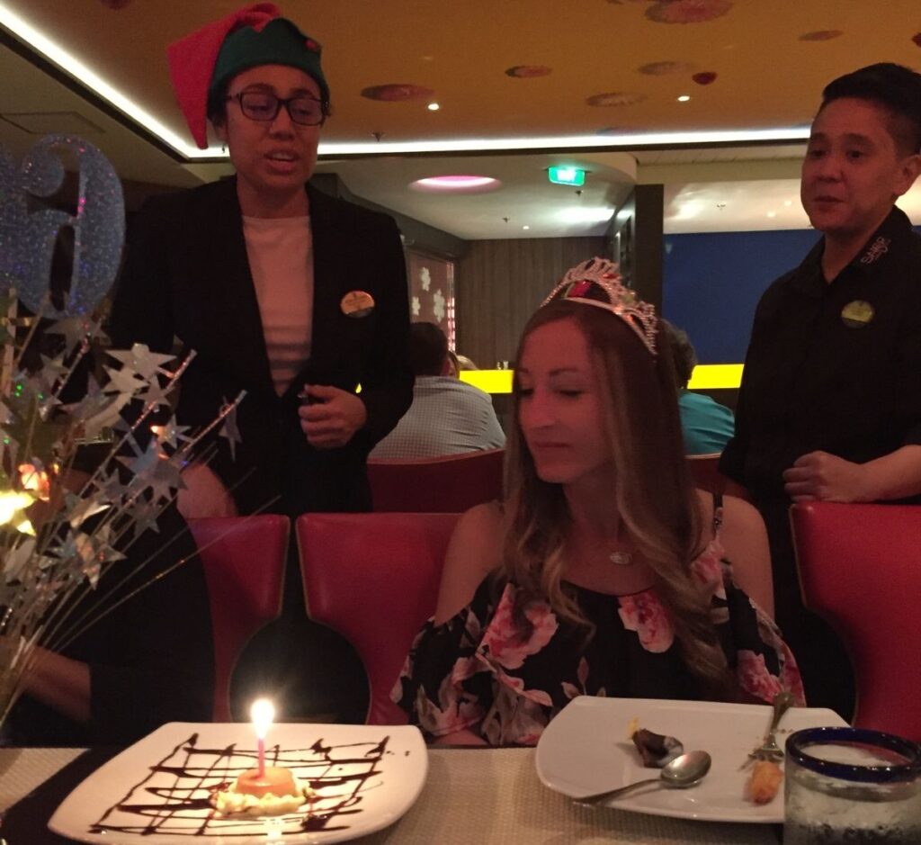 A woman wearing a tiara sits at a cruise ship dining table in front of a birthday dessert with a lit candle, while two crew members stand beside her.