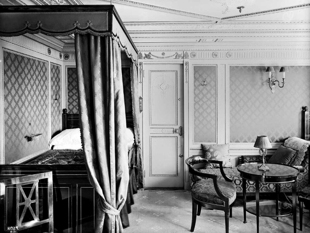 Black-and-white photograph of a Titanic first-class suite with a curtained bed, seating area, and ornate Edwardian decor.