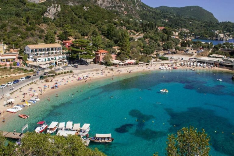 Aerial view of a sandy beach and clear turquoise water in Corfu, Greece, with small boats moored near the shore and green hills rising behind the bay.