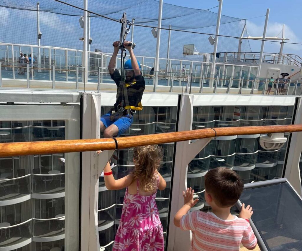 A man rides a zip line across the upper deck of a cruise ship while two children watch from behind a railing, with sports courts and ship balconies in the background.