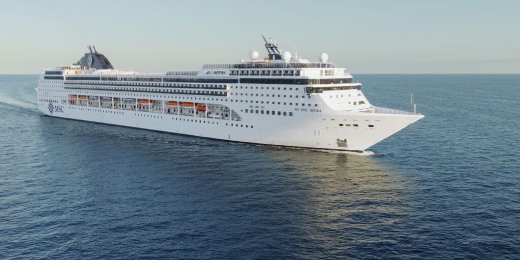 The MSC Opera cruise ship is captured cruising the open sea, featuring its distinctive navy blue and white exterior, multiple decks with lifeboats, and a sleek design, set against a backdrop of clear skies and calm waters.