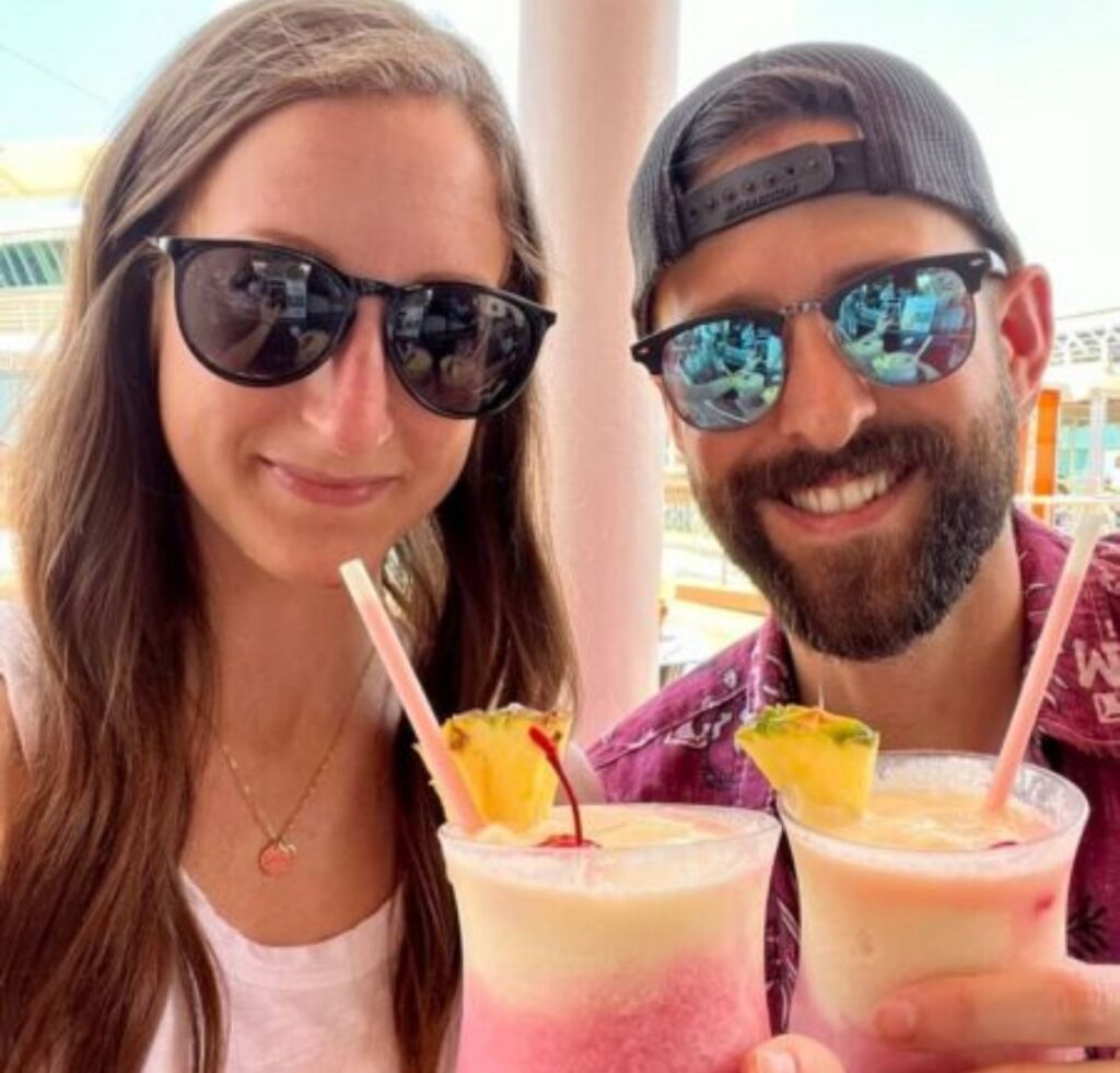 A smiling couple wearing sunglasses holds tropical frozen drinks garnished with pineapple while relaxing outdoors on a cruise ship.