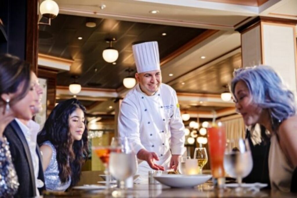 Cruise ship chef serving dinner to guests seated at a specialty dining restaurant.
