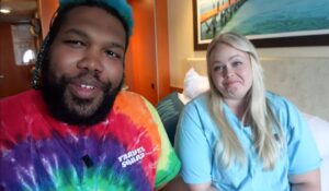 Two travel YouTubers sit in a cruise cabin filming a video update, facing the camera. One wears a colorful tie-dye shirt, while the other wears a light blue top.