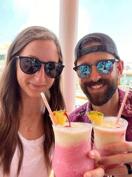 Two adults wearing sunglasses on a cruise ship deck, smiling and holding frozen tropical drinks garnished with pineapple.