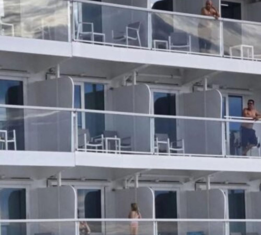 Rows of cruise ship balcony cabins with passengers visible on several balconies, each furnished with chairs and small tables.