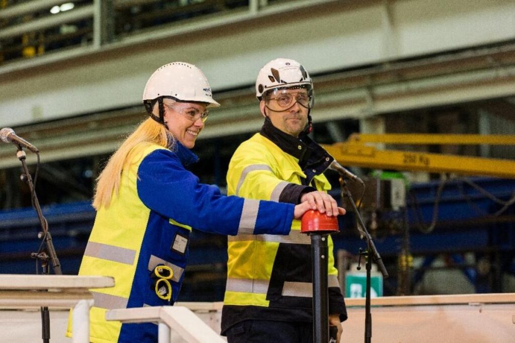 Two shipyard workers in hard hats and high-visibility jackets press a red button during a cruise ship construction ceremony.
