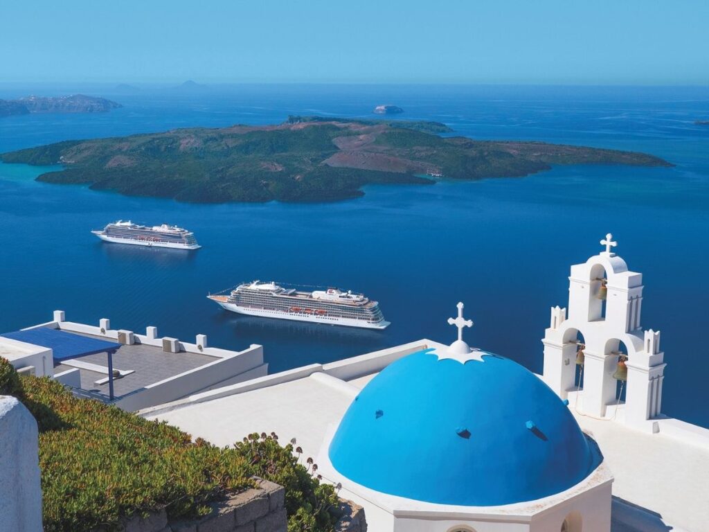 Two Viking cruise ships sail off Santorini, seen beyond whitewashed buildings and a blue church dome overlooking the sea and a Greek island in the background.