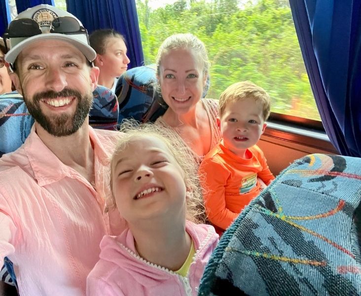 Family smiling for a selfie while seated on a tour bus, with two adults and young children by a window overlooking greenery outside.