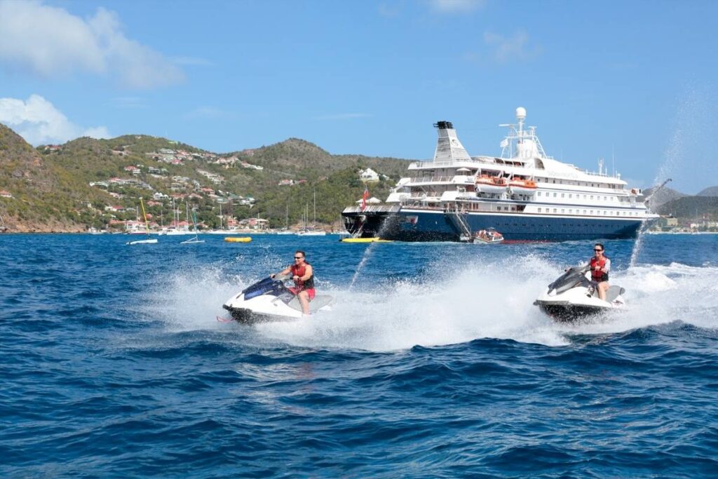 SeaDream yacht anchored off a Caribbean island as two guests ride jet skis across the water in front of the ship.
