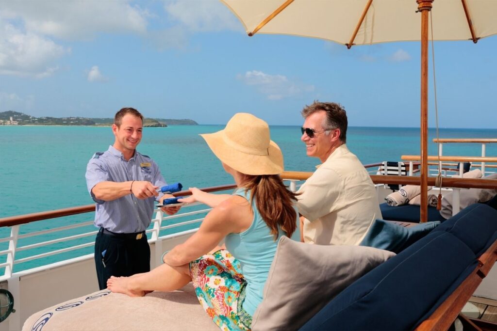 SeaDream crew member serves two guests relaxing on an outdoor deck overlooking turquoise Caribbean waters.