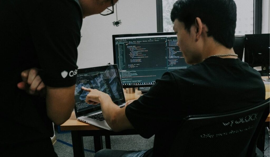Two people reviewing computer code on laptop and desktop screens during a cybersecurity investigation.