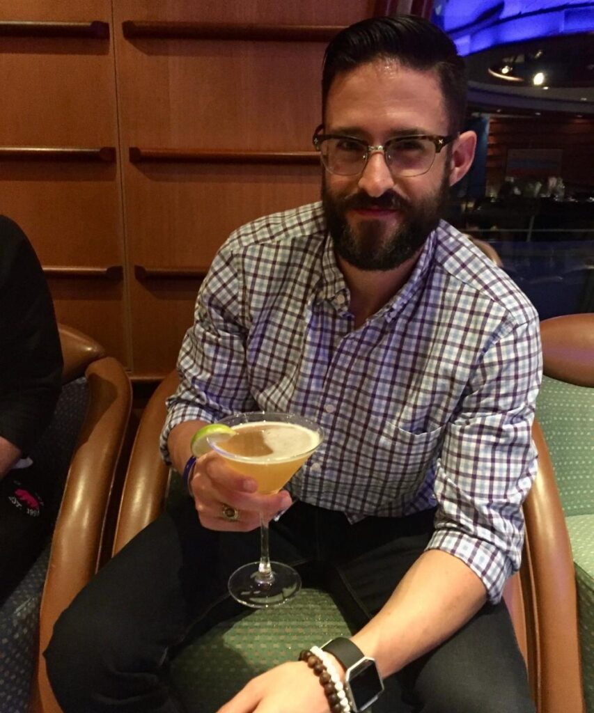 Man sitting in a lounge on a cruise ship holding a cocktail in a martini glass, with wood paneling and seating around him.