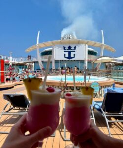 Two frozen tropical cocktails held up on the pool deck of a Royal Caribbean cruise ship, with the Royal Caribbean logo, hot tubs, loungers, and blue sky in the background.