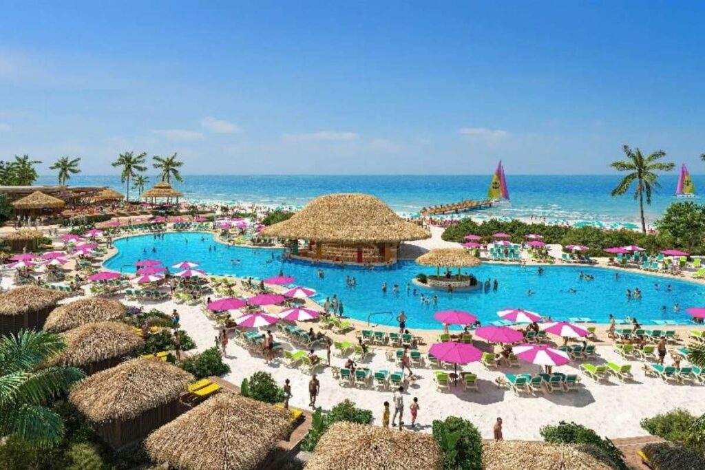 Royal Beach Club Cozumel pool area with lounge chairs, pink umbrellas, thatched-roof structures and the ocean in the background.