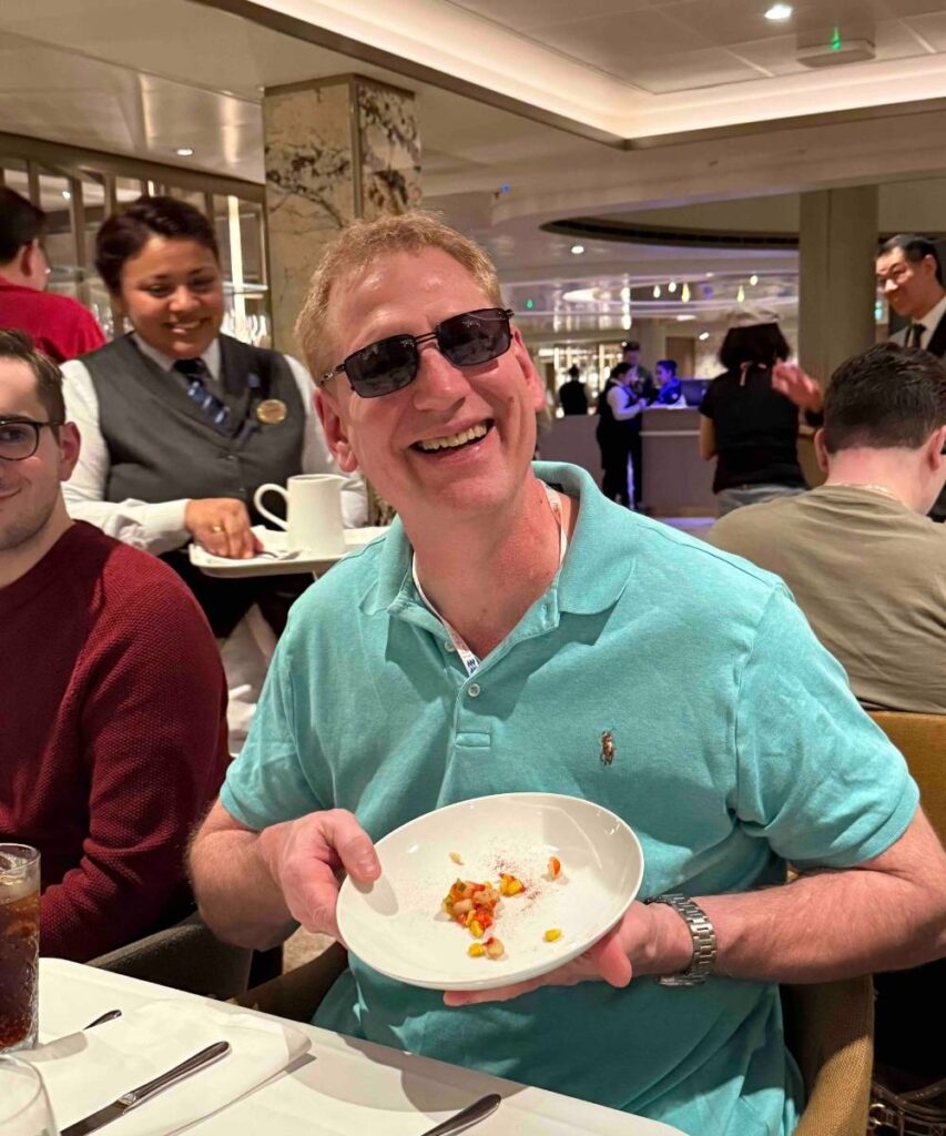 Smiling cruise passenger seated at a dining table onboard a ship while holding a plate of food.